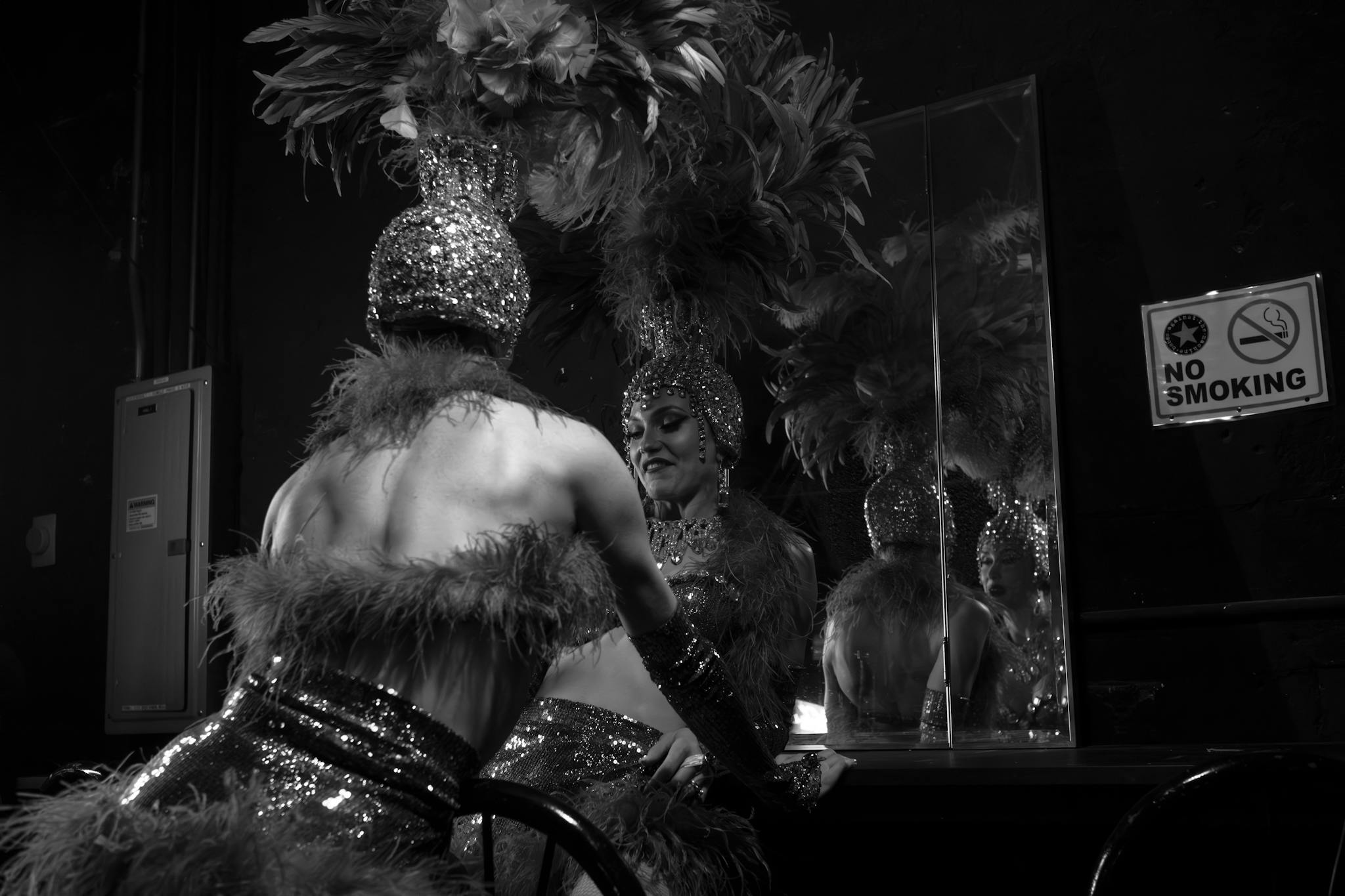 Dancer in feathered headdress and sequined costume gets ready backstage, reflecting in mirror.